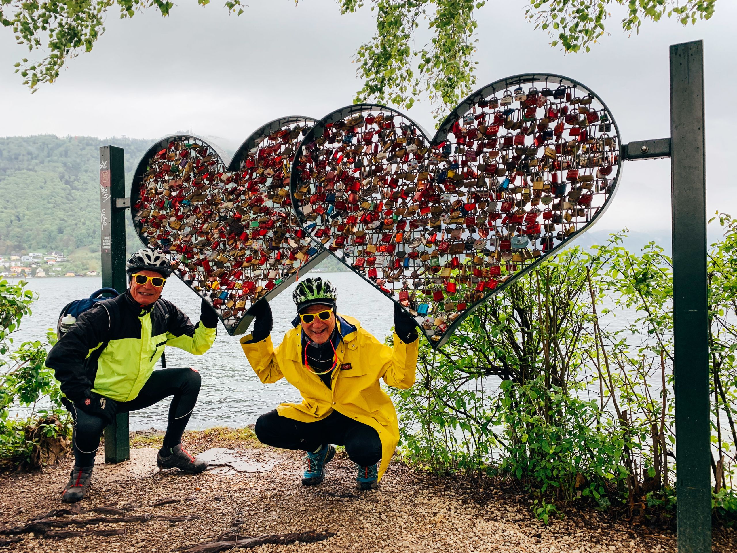 Coppia di ciclisti in posa di fronte a recinzione a forma di cuore piena di lucchetti lungo la Ciclabile del Salzkammergut